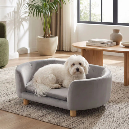 Canapé gris pour chiot et chien, structure en bois de bouleau , dossier haut et entourant à 360° au design moderne et arrondi avec son coussin en éponge 100% coton haute densité et ergonomique déhoussable et lavable en machine, housse en velours douce et résistante, sur pied en bois massif pour chien de petite taille- un petit chien blanc à poils longs couché confortablement sur le canapé ergonomique au design moderne de couleur gris au pied du lit de ses maitres dans le salon lifestyle de l'appartement