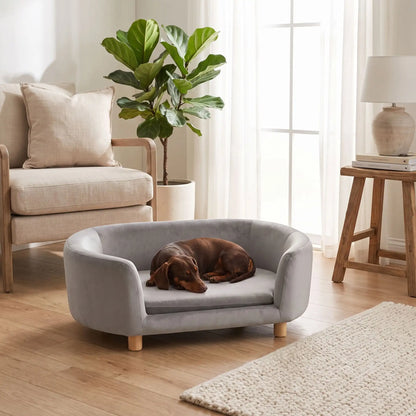 Canapé gris pour chiot et chien, structure en bois de bouleau , dossier haut et entourant à 360° au design moderne et arrondi avec son coussin en éponge 100% coton haute densité et ergonomique déhoussable et lavable en machine, housse en velours douce et résistante, sur pied en bois massif pour chien de petite taille- un chien de race miniature qui dort couché confortablement en boule sur le canapé ergonomique au design moderne de couleur gris au pied du canapé de ses maitres dans le salon de l'appartement