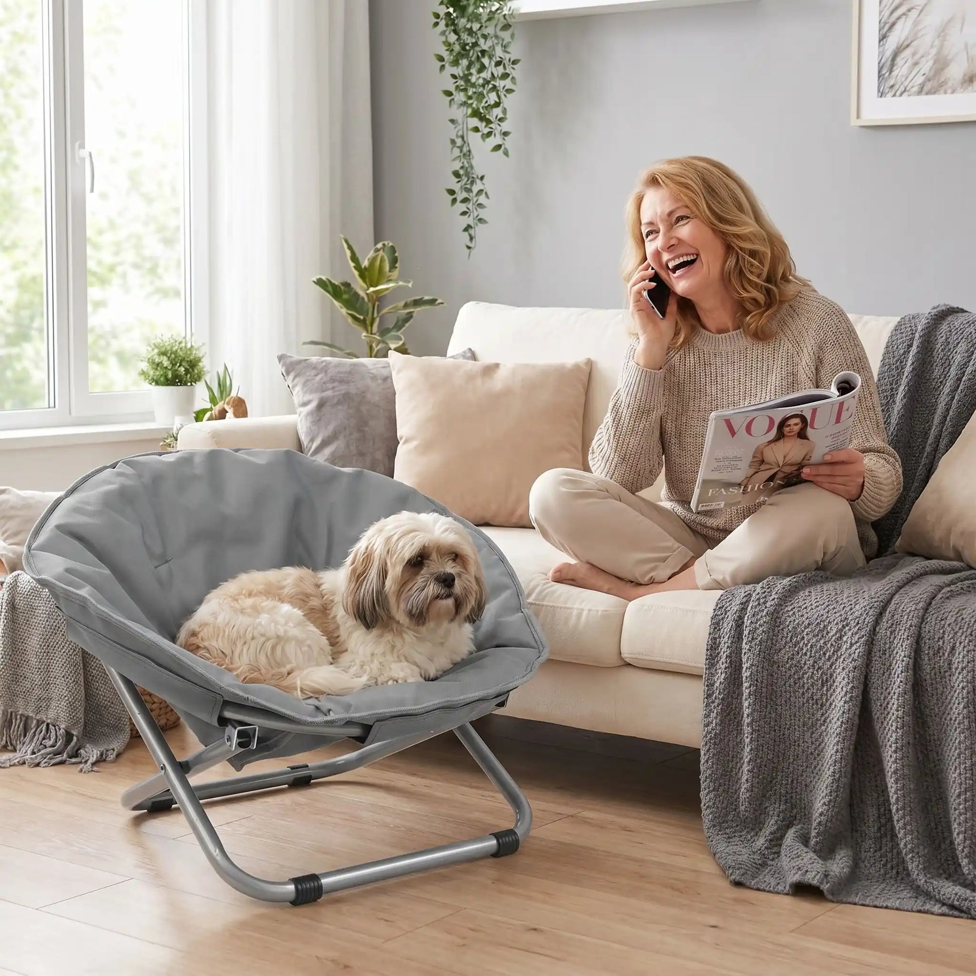 Chaise-pour-chien-chiot déhoussable et lavable, imperméable, structure design en acier robuste arrondie comme un cocon entourant avec deux hauteurs adaptables, pliable avec sa housse en nylon résistant aux griffes pour chiot et chien de 0 à 7 kilos-un chien de petite raille à long poils couché dans la chaise entourante sur pieds de couleur grise à côté de sa maitresse qui rit au téléphone avec son magazine dans le salon de l'appartement