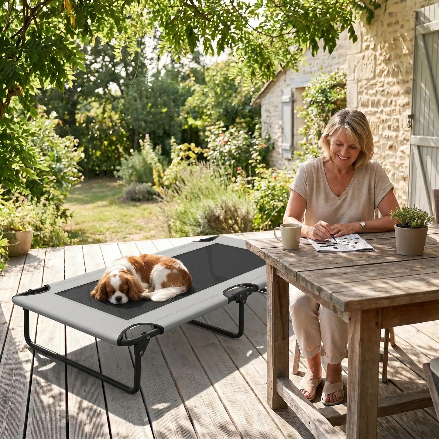 Hamac pour chien de 107 cm, en tissu oxford 100% imperméable et maille respirante et structure en acier robuste, polyvalent pour une utilisation interieur et exterieur, surélevé avec 3 barres transversales et patin anti rayure pour une stabilité pour chiot et chien de petite à moyenne taille jusqu'à 30 kilos-un chien à poils long et épais qui dort couché au frais sur son lit surélevé gris foncé au pied de sa maitresse qui joue aux mots croisée sur la table en bois de la terrasse de la maison dans le sud 