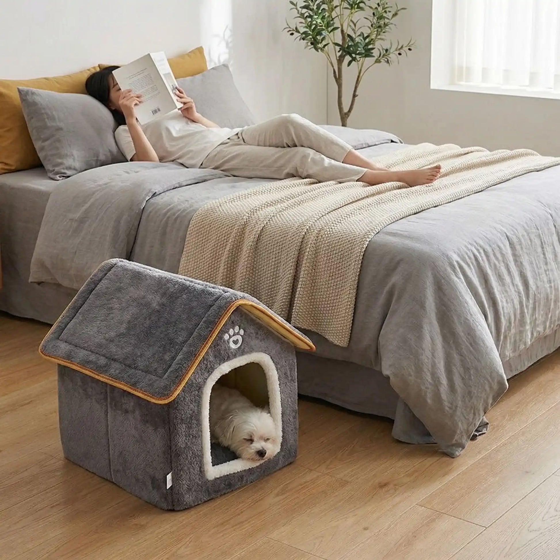 Maison-chien-intérieure-chiot-mini, pliable, légère et transportable, avec matériau isolant efficace contre le bruit et du froid, niche antidérapante, lavable en machine avec toit amovible, de couleur grise pour chiot et chien de 0 à 5 kg, disponible en taille XS et S- un chien de petite taille qui dort paisiblement dans la petite niche intérieure sur le sol de la chambre à côté du lit de sa maitresse allongé qui lit son livre tranquillement