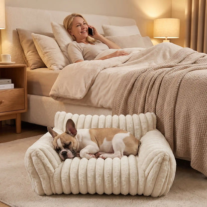 Mini-canapé-chien-chiot-déhoussable, lavable en machine ,coton, housse en peluche douce au toucher velours, rembourrage ergonomique en mousse à mémoire de forme pour un soutien optimal, disponible en  couleurs blanc et rose et 3 tailles S, M et L pour chiot et chien de petite taille de 0 à 10 iols max-un chiot de race moyenne qui dort paisiblement niché entre les deux accoudoirs rembourré du mini fauteuil pour chien de couleur blanc crème au pied du lit de sa maitresse allongée au téléphone 