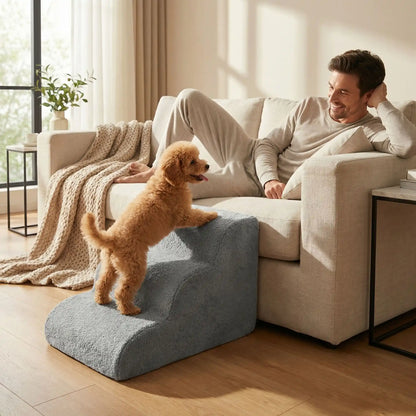 Petit-escalier-pour-chien-anti-deformationen mousse éponge haute résilience qui atténue la pressions sur  ses articulations, résistant et doux pour ses coussinets, déhoussable et lavable en machine de couleur gris pour chien de petite taille, polyvalent compact et transportable-un caniche qui monte sur l'escalier pour rejoindre son maitre qui le regarde en souriant allongé sur le canapé du salon de l'appartement
